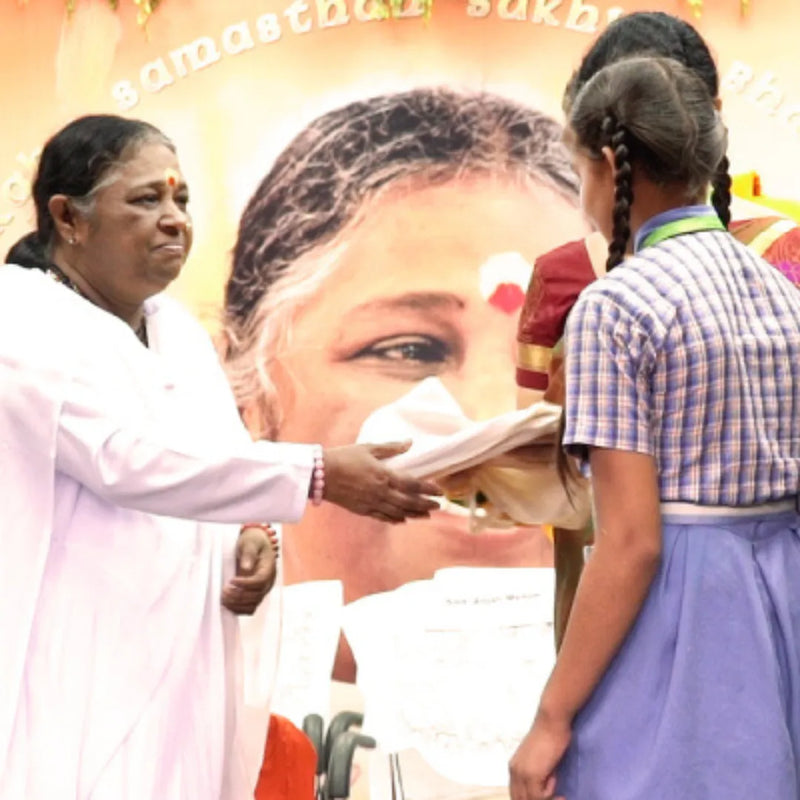 Sri Mata Amritanandamayi Devi (Amma) presenting a ceremonial gift to a young student during an event, symbolizing compassion and support for education. The image reflects Amma's role in promoting humanitarian values and nurturing future generations.
