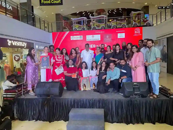 A large group of people posing on a stage in a shopping mall auditorium or event space, with a red backdrop, speakers, and a black draped floor covering. Several people are holding framed certificates or awards.