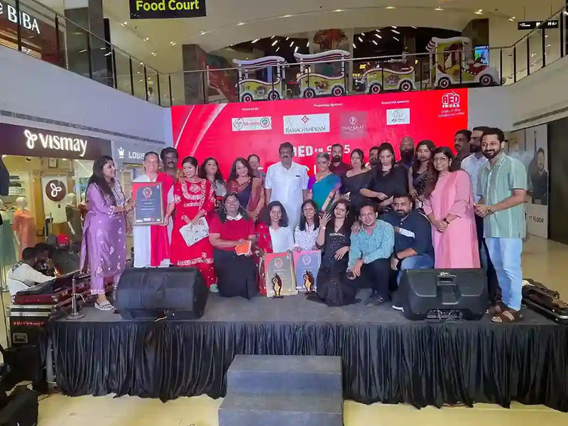 A large group of people posing on a stage in a shopping mall auditorium or event space, with a red backdrop, speakers, and a black draped floor covering. Several people are holding framed certificates or awards.